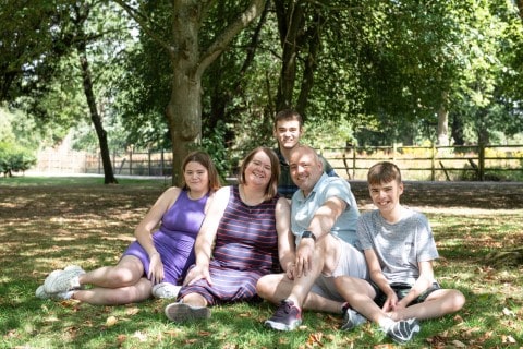 Family of five smiling and sitting on grass under trees during a relaxed outdoor photography session – natural family portrait by LB Photography