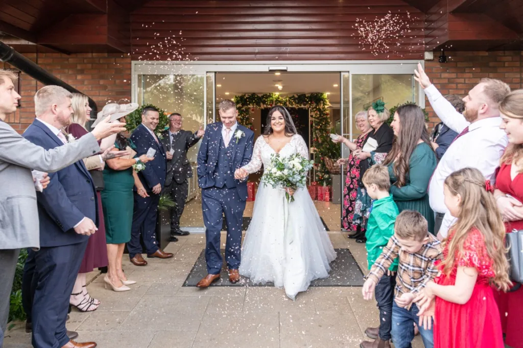 Guests cheering and throwing confetti as Leah and Ryan walk hand-in-hand outside Draycote Hotel during their winter wedding