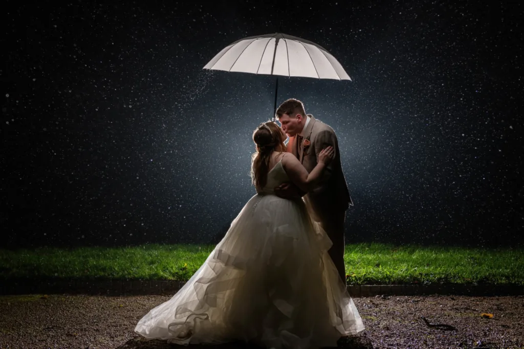 Bride and groom kissing under a white umbrella at night, surrounded by raindrops — a romantic example of rain on your wedding day photos captured by LB Photography.