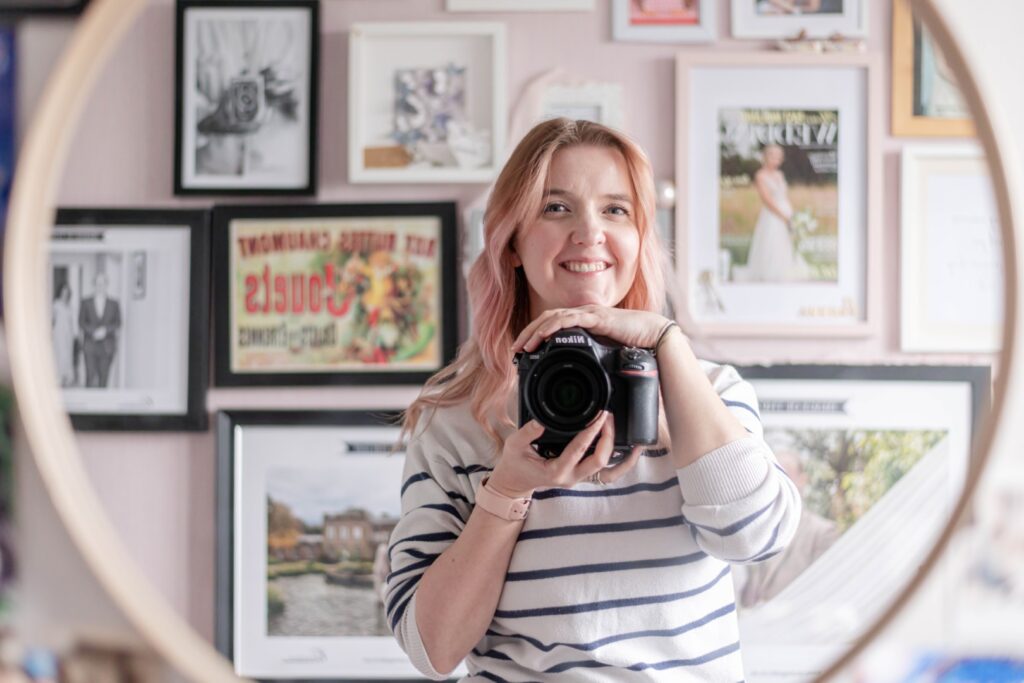 A woman with pink hair smiles while holding a Nikon camera, surrounded by framed artworks and photographs in a cozy interior setting.