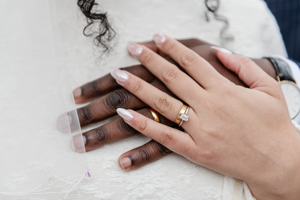Close-up of intertwined hands showcasing wedding rings, with a delicate veil in the background.