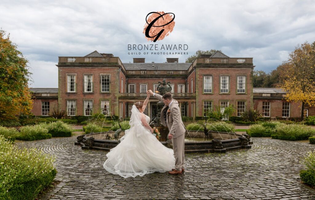 A bride and groom share a joyful dance in front of a historic mansion, surrounded by lush greenery and a fountain, on an overcast day.