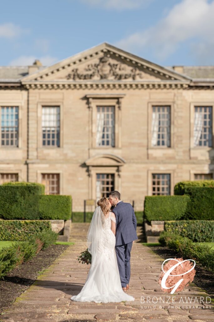 A couple embraces in wedding attire, walking hand in hand in front of a grand stone building surrounded by neatly trimmed hedges and lush greenery.