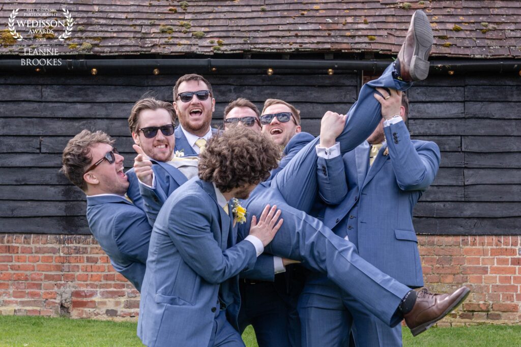A group of well-dressed groomsmen in blue suits and sunglasses joyfully lifting one of their friends in a playful manner, set against a rustic wooden backdrop.