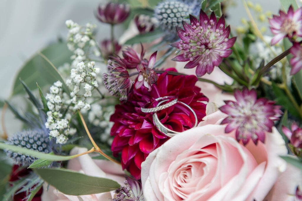 Close-up of intertwined wedding rings resting on vibrant flowers, showcasing a romantic floral bouquet with pink roses and assorted blooms.