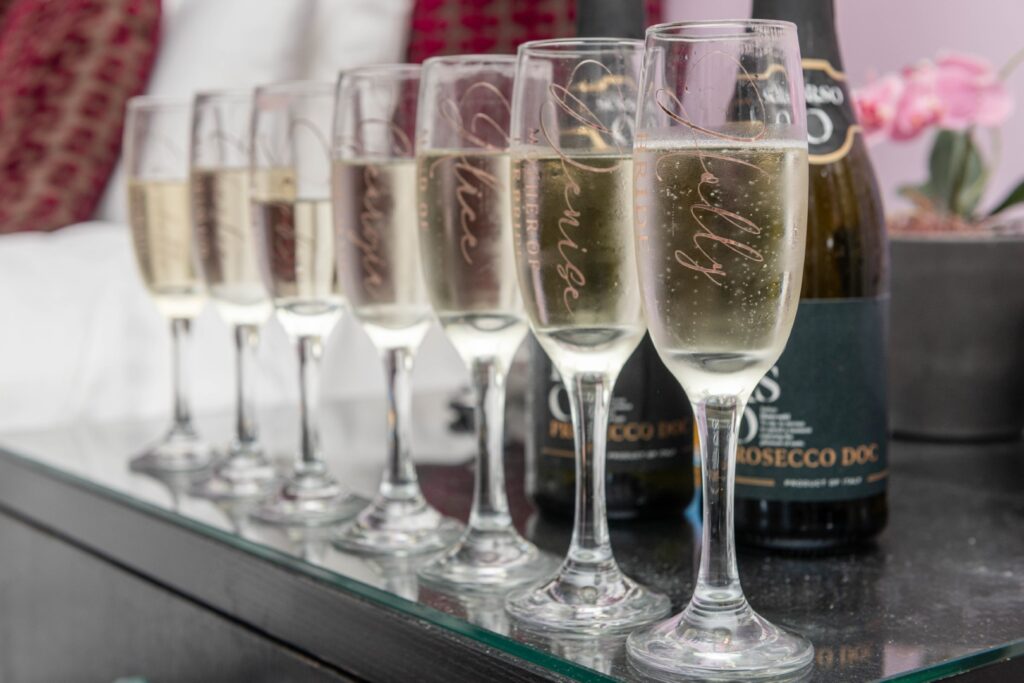 A row of customized champagne flutes filled with sparkling wine, accompanied by bottles of Prosecco on a glass table.
