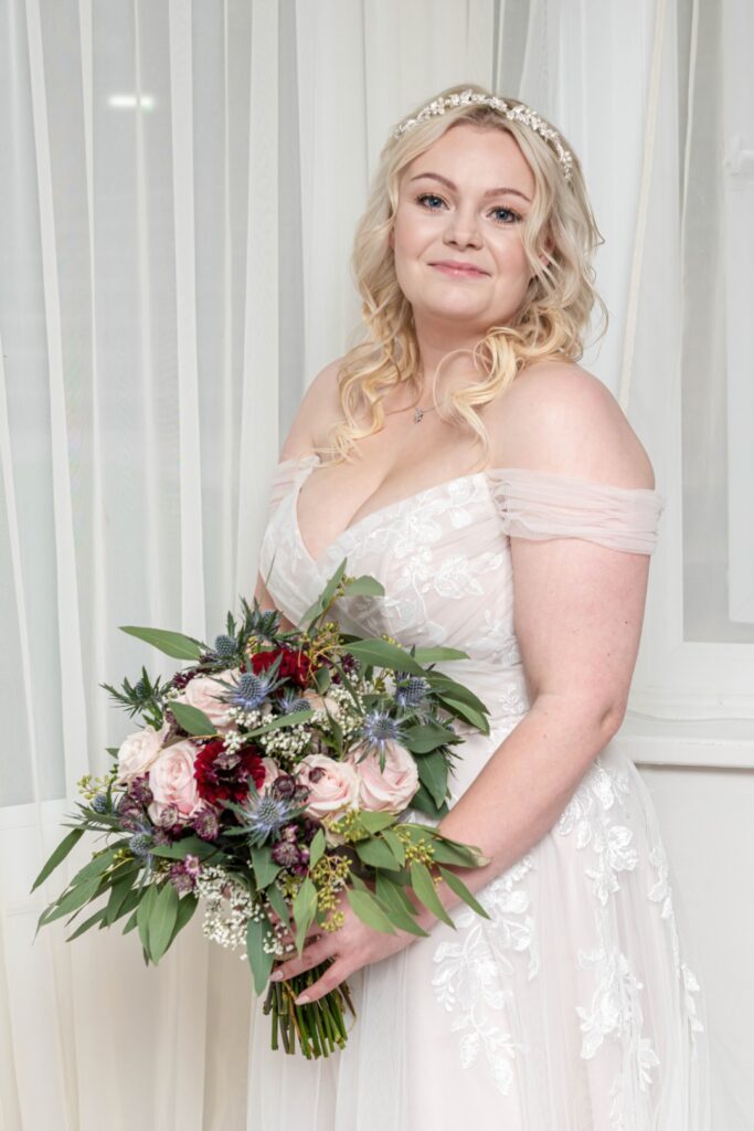 A bride in an elegant off-the-shoulder gown holds a beautiful bouquet while standing in front of sheer curtains.