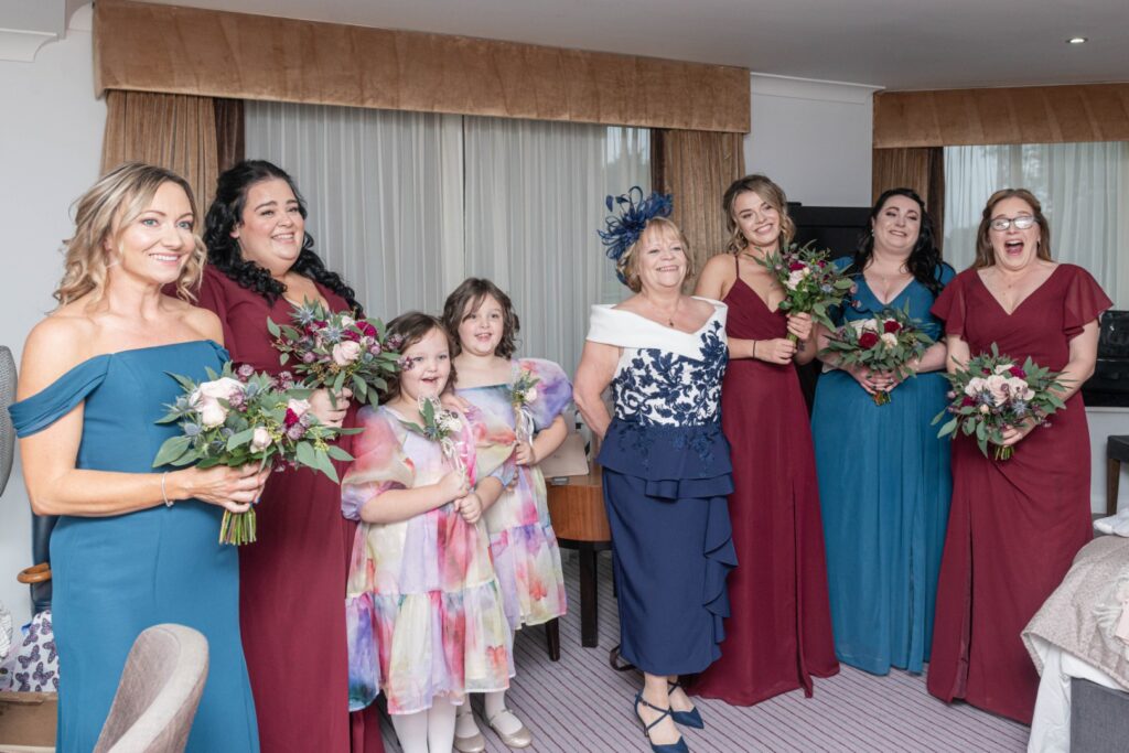 A joyful bridal party consisting of women in elegant dresses and two young girls, all holding bouquets, posing inside a beautifully decorated room.
