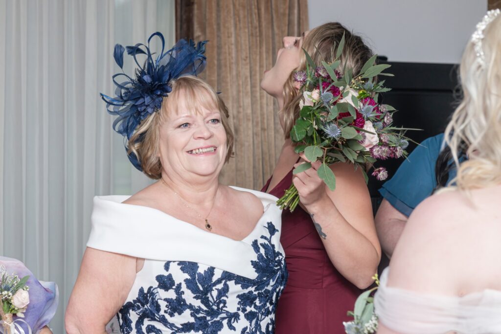 A smiling woman in an elegant dress and hat poses for a photo while another woman with a bouquet laughs in the background.