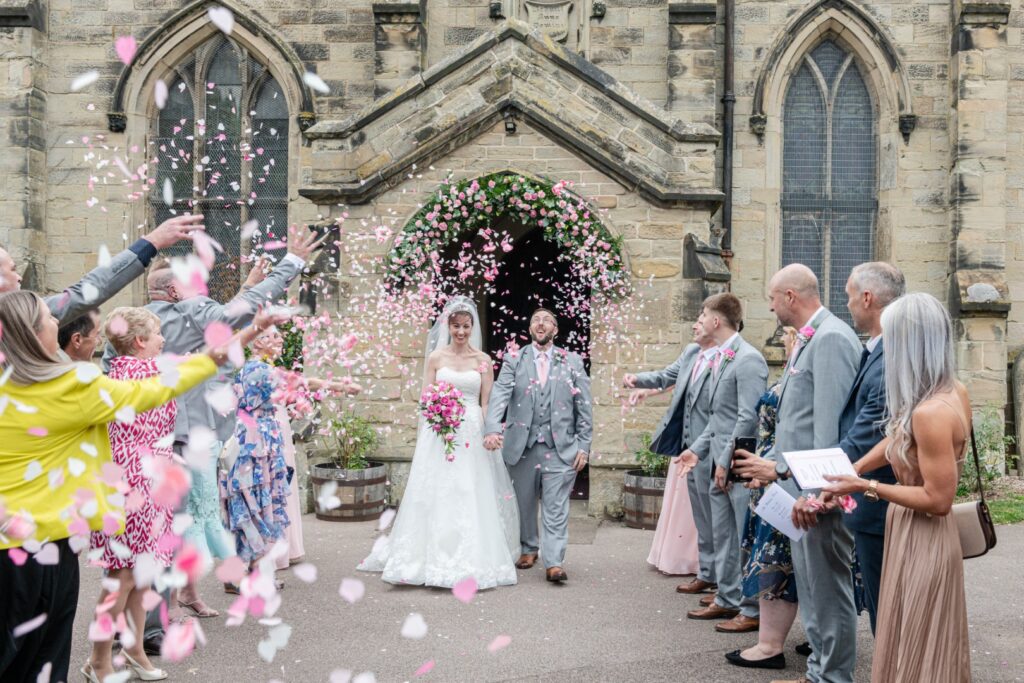 A joyful couple exits the church surrounded by guests throwing pink and white petals, capturing a moment of happiness and celebration.