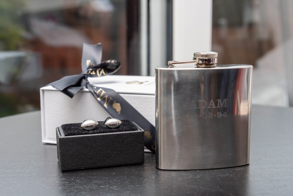 A stainless steel flask engraved with "ADAM 19-02-94" next to a black box containing silver cufflinks, all on a dark surface with a gift box and ribbon in the background.