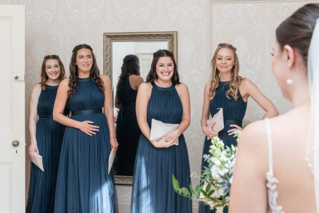 Bridesmaids' Excitement::A group of bridesmaids in navy dresses smiles at the bride, capturing a moment of joy and anticipation before the wedding ceremony.