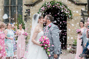 Wedding Celebration Kiss:::A joyful couple shares a kiss amidst a flurry of confetti, surrounded by friends and family during their wedding ceremony outside a beautiful stone church.