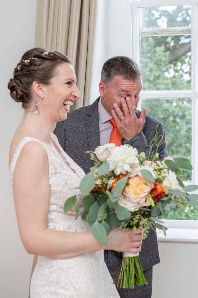 A bride laughing joyfully while holding a bouquet, with a male figure smiling and wiping his eyes in the background.