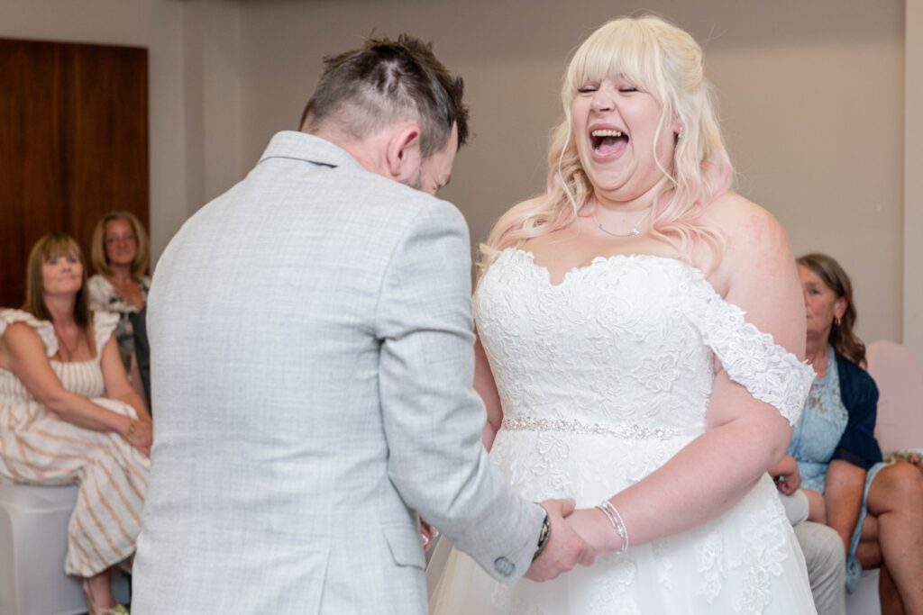 A bride laughs joyfully while holding hands with her partner during their wedding ceremony, surrounded by smiling guests in the background.