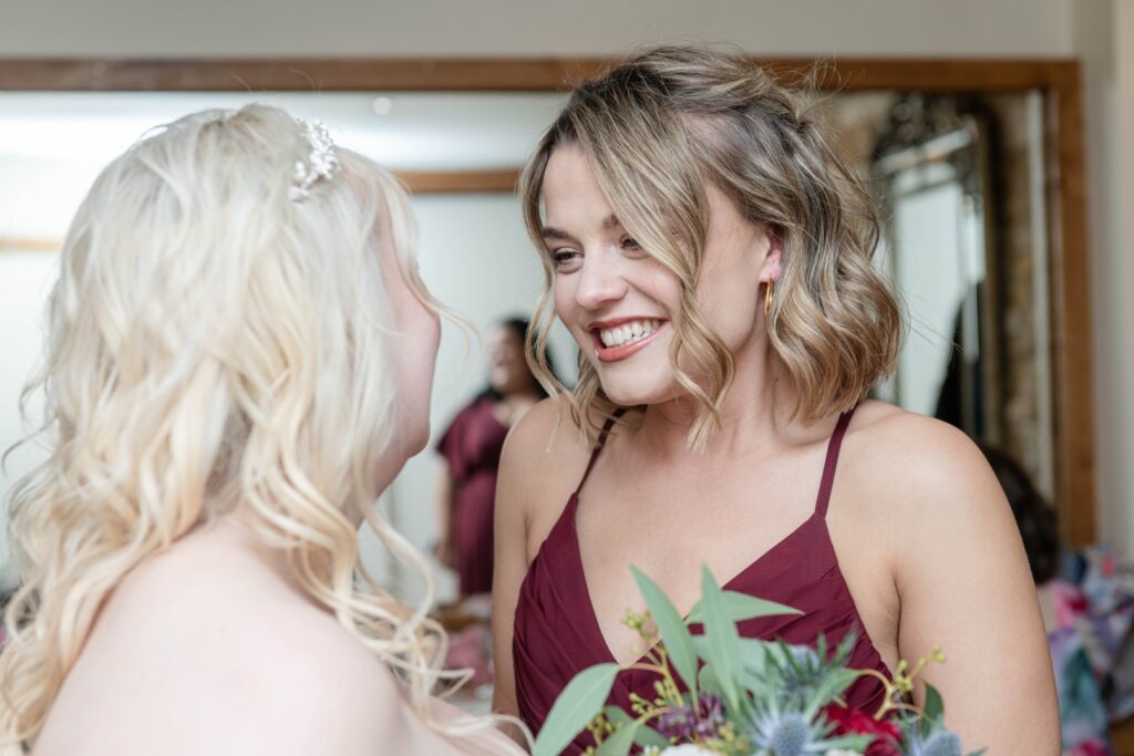 Two bridesmaids share a heartfelt moment, smiling and enjoying each other's company, with a floral arrangement visible in the foreground.