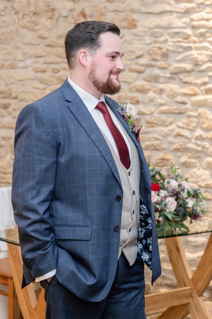A well-dressed man in a blue checkered suit with a maroon tie and floral boutonniere, standing beside a floral arrangement against a stone wall backdrop.