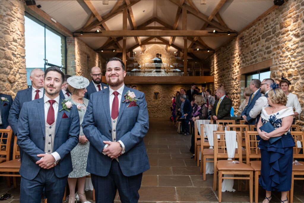 Two groomsmen in blue suits stand at the front of a beautiful wedding venue, while guests are seated in the background, creating an elegant atmosphere.