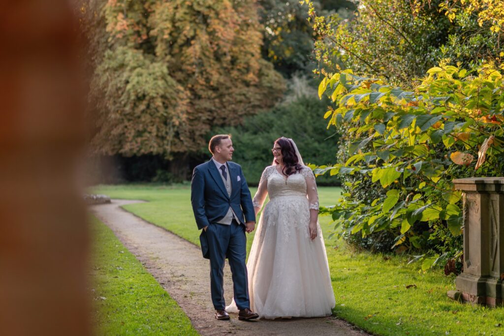 A couple dressed in wedding attire walking hand-in-hand along a scenic pathway surrounded by lush greenery and autumn foliage.