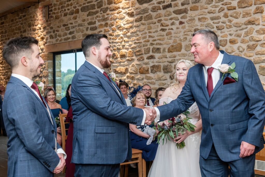 Two groomsmen shaking hands in front of a bride during a wedding ceremony, with guests smiling in the background amidst a rustic stone setting.