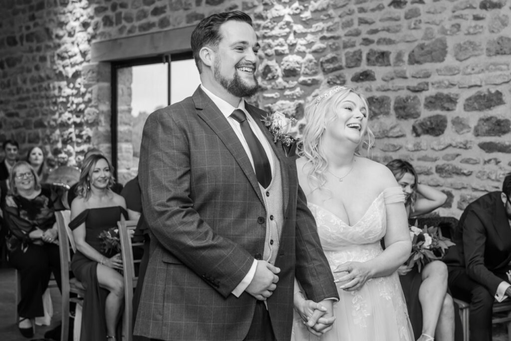 A bride and groom smile at each other during their wedding ceremony, surrounded by seated guests in a rustic venue.
