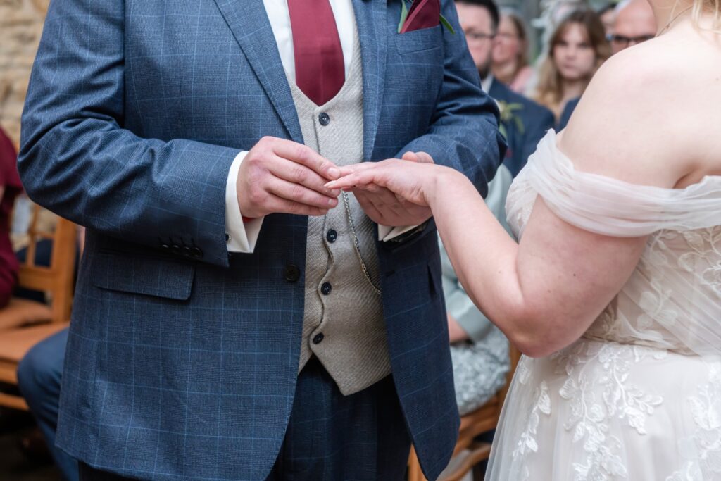 A groom placing a wedding ring on the bride's hand during their ceremony, surrounded by guests in the background.