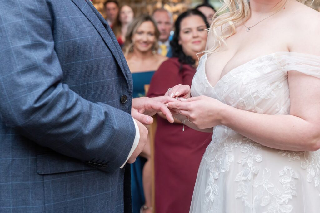 A bride places a ring on her groom's finger during a wedding ceremony, surrounded by guests in a rustic venue.
