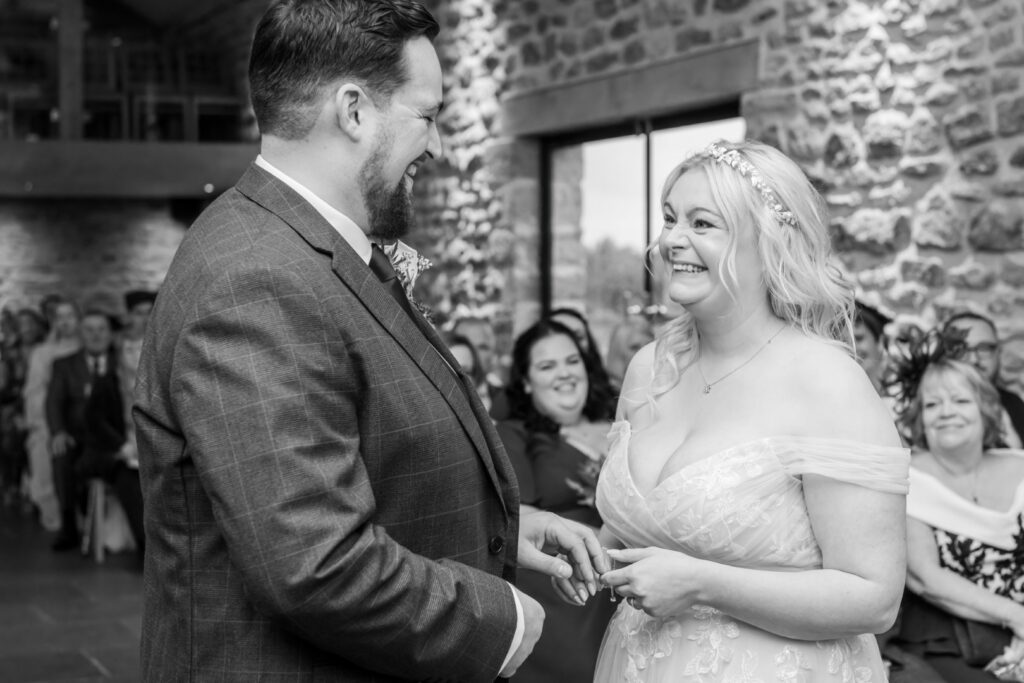 A couple exchanges wedding vows, smiling at each other surrounded by family and friends in a stone-walled venue.