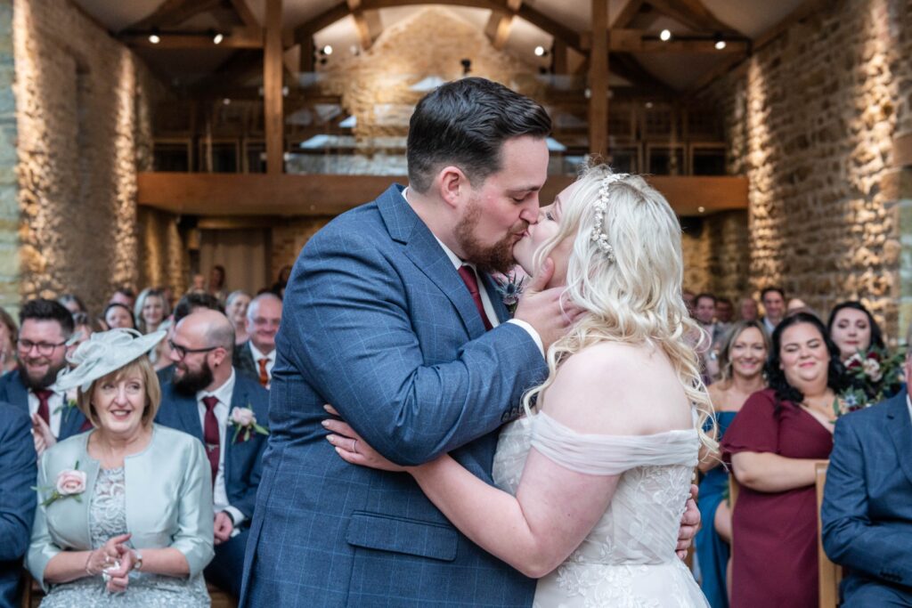 A couple sharing a romantic kiss during their wedding ceremony at Dodford Manor, surrounded by a joyful audience in a beautifully decorated venue.