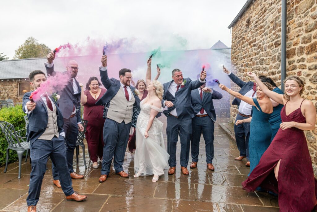A joyful wedding party celebrating with colorful smoke bombs in Dodford Manor, showcasing laughter and festive attire.