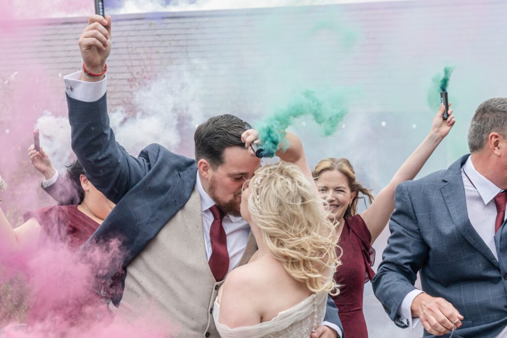 A couple shares a romantic kiss while surrounded by guests holding colourful smoke bombs, creating a vibrant and joyful atmosphere. at Dodford Manor