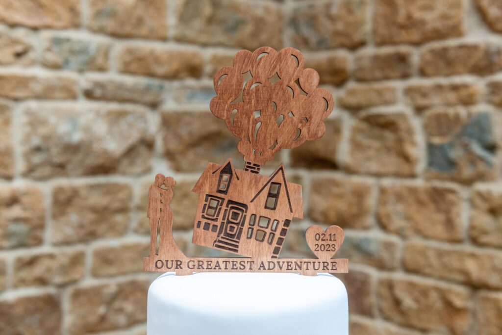 A wooden cake topper featuring a house with balloons, a couple, and the date 02.11.2023, set against a textured stone background.
