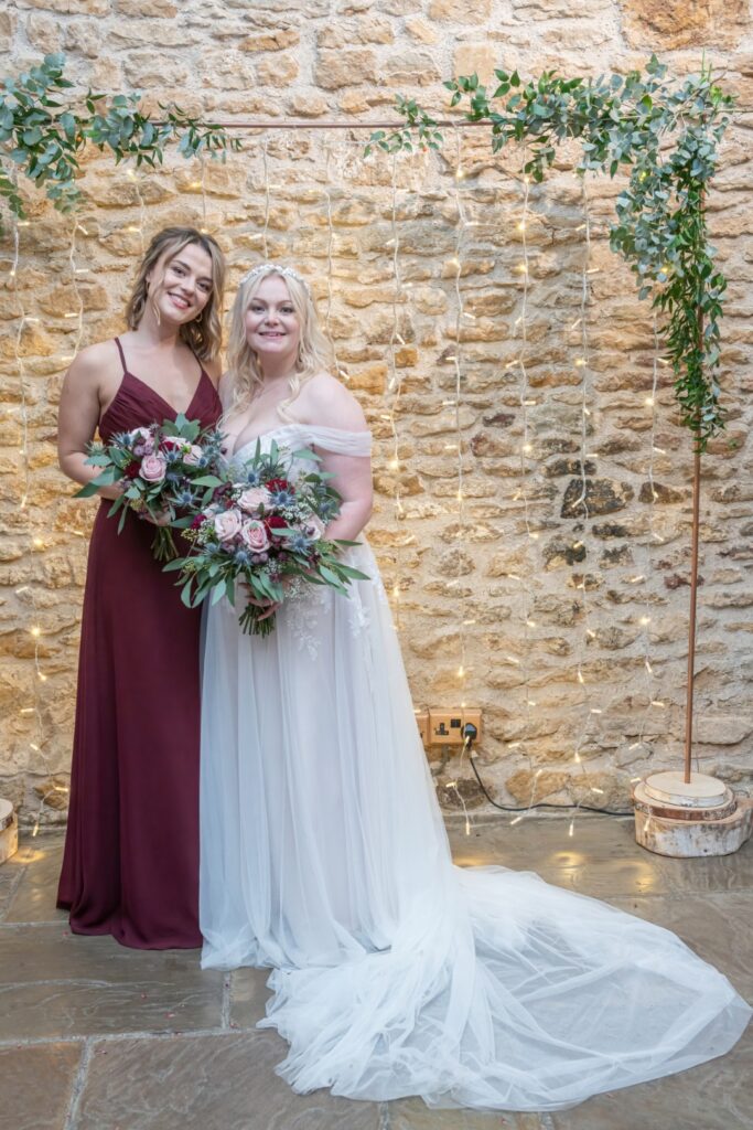 Two women in formal dresses pose together with floral bouquets in front of a rustic stone wall adorned with fairy lights at dodford manor