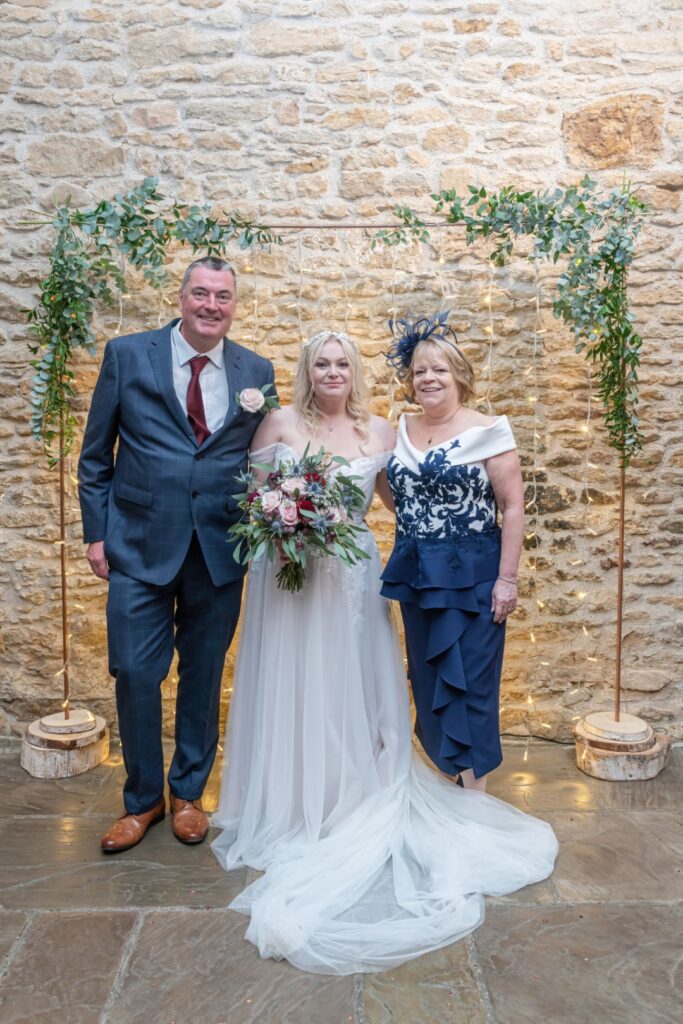 A bride poses with her parents in a charming venue featuring stone walls and soft lighting inside of Dodford manor