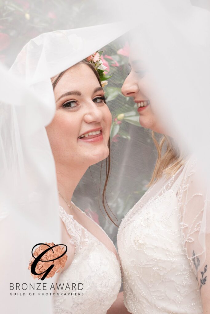 A close-up of two brides smiling warmly at each other, framed by a delicate veil with a floral backdrop, exuding joy and love.