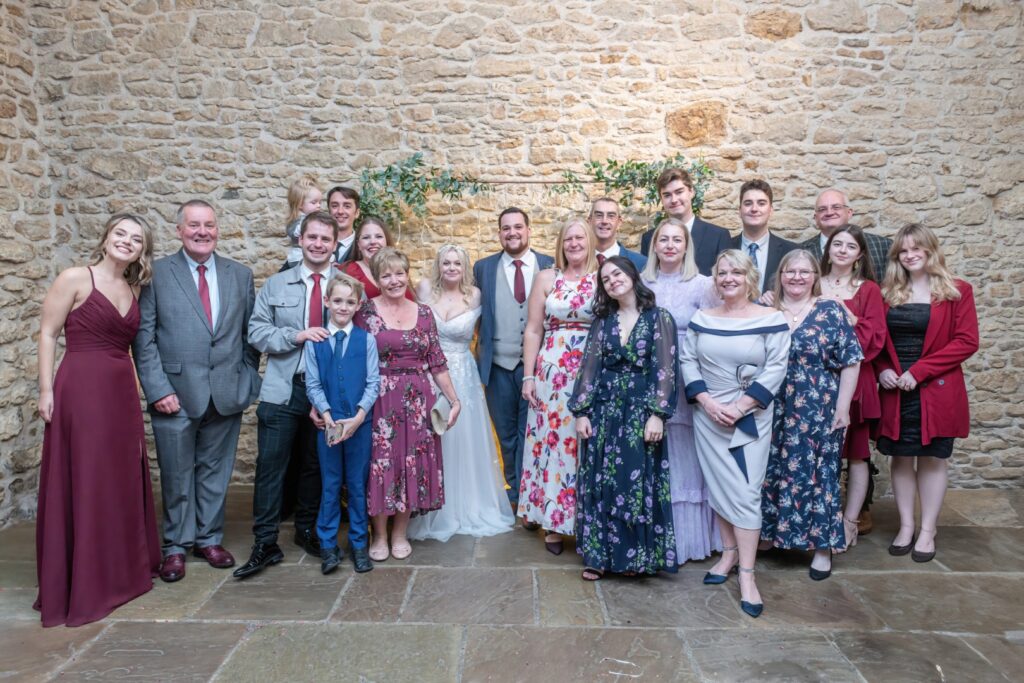 A large group of people dressed in formal attire posing together, smiling in front of a stone wall adorned with greenery, capturing a joyful family moment.