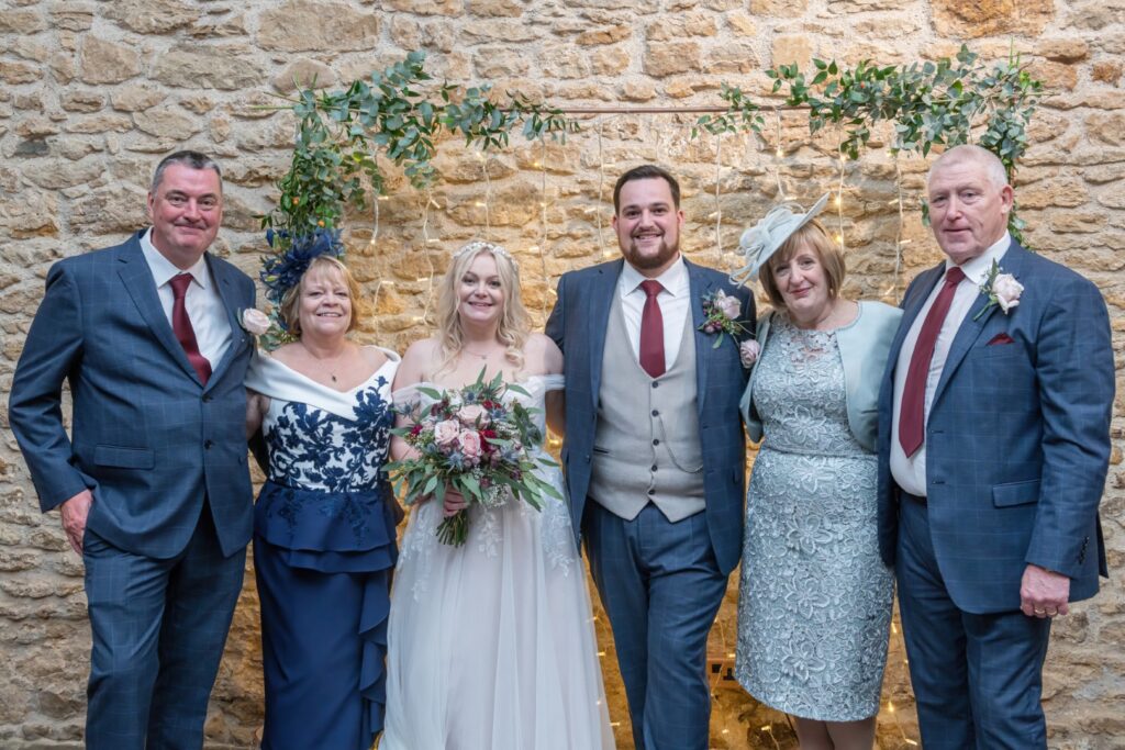 A joyful group photo of a bride in a white dress and her family, standing in front of a decorated stone wall with greenery and fairy lights.