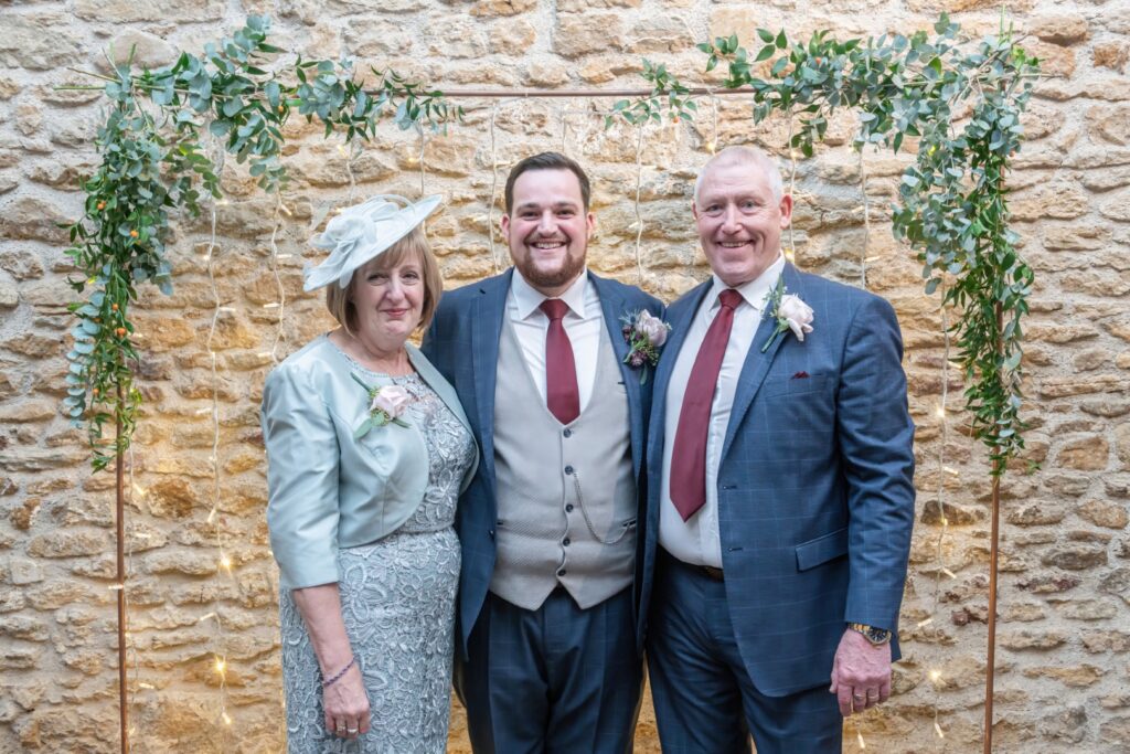 A smiling man stands between his parents, who are dressed elegantly, with a decorative backdrop of greenery and fairy lights.