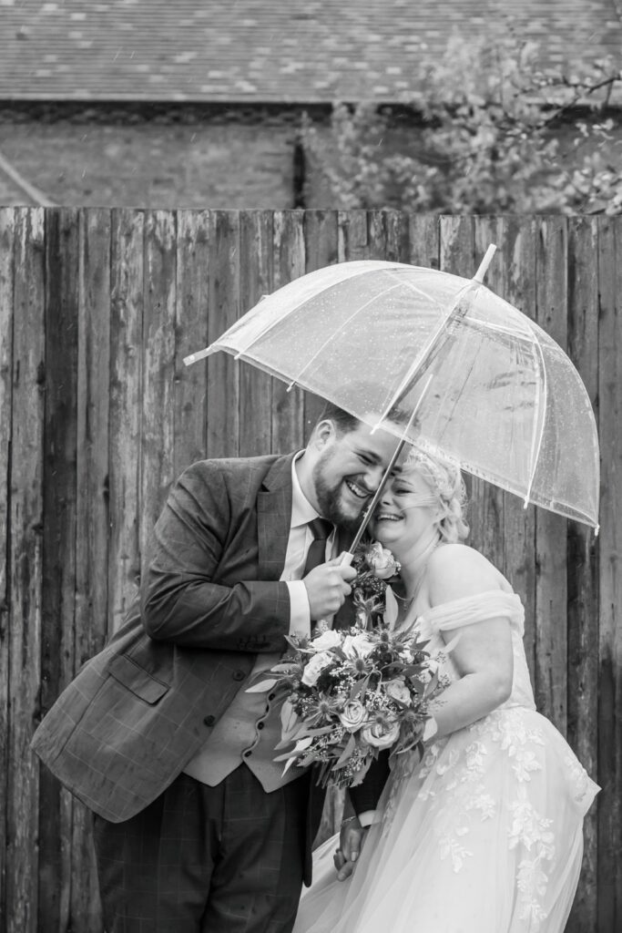 A happy couple embraces under a clear umbrella, sharing a moment of love and laughter amidst a rainy backdrop, with the bride holding a bouquet of flowers.