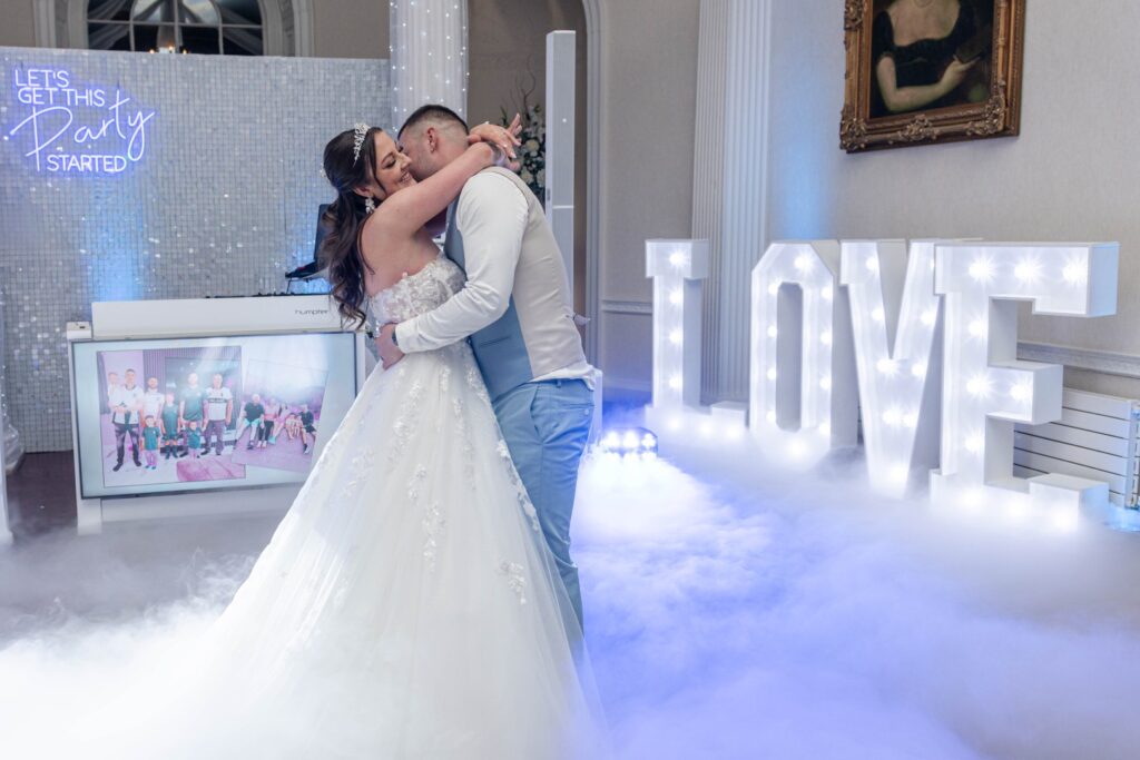 A couple embraces in a romantic moment during their wedding reception, surrounded by decorative lights and fog.