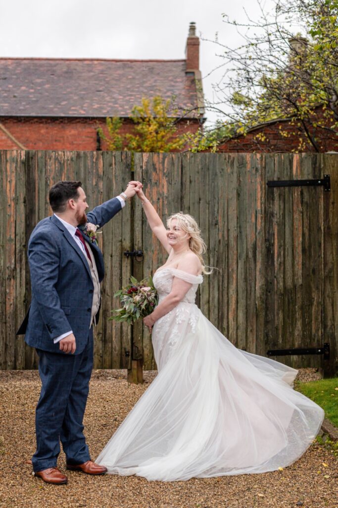 A joyful couple dances together outdoors, embracing the moment of their wedding with a scenic backdrop of trees and an old wooden fence.