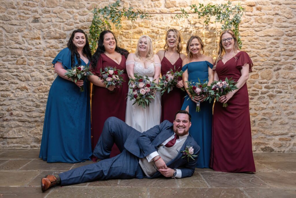 A joyful wedding party composed of the bride and bridesmaids, proudly posing with bouquets in a picturesque setting. The groom is playfully lying on the ground in front of them Dodford Manor