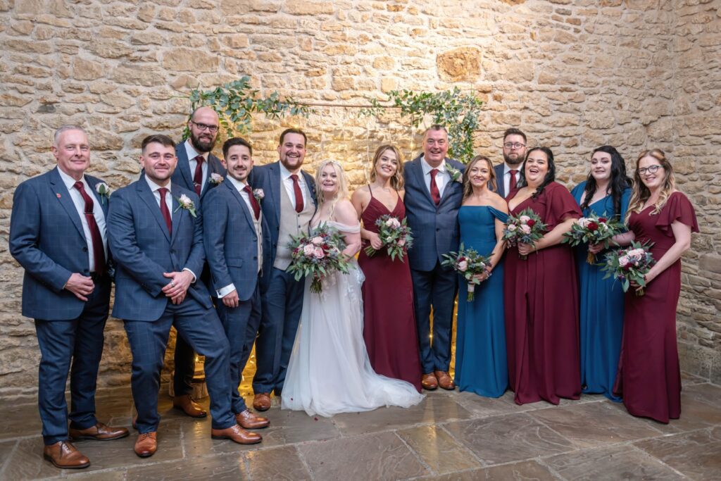 A joyful wedding party poses together in a beautifully decorated venue, showcasing a mix of elegant attire and floral arrangements.