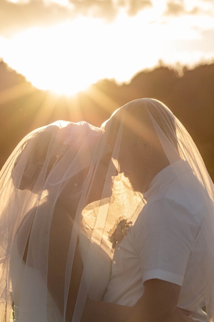 A close-up of a bride and groom sharing a tender moment under a veil, illuminated by the sun setting behind them.