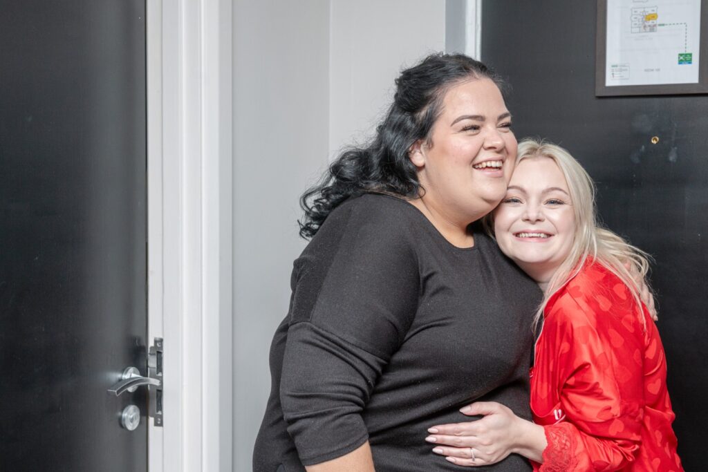 Two women sharing a joyful embrace, smiling widely in a doorway with a black backdrop.