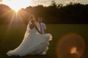 A couple dances together on a grassy field during sunset, capturing a romantic moment with sunlight illuminating their joyful expressions.
