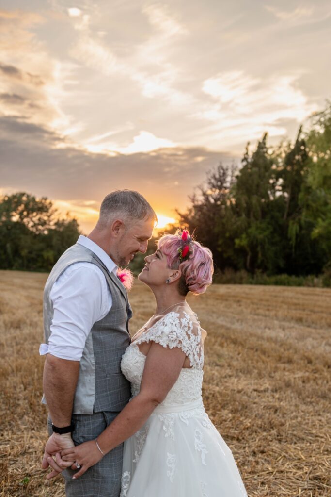 A couple stands close together in a field during sunset, gazing into each other's eyes with smiles, with the woman wearing a white wedding dress and the man in a gray suit.