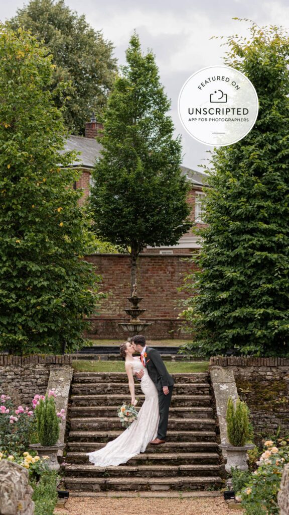 Bride and groom sharing a kiss on stone steps surrounded by lush greenery and blooming flowers.