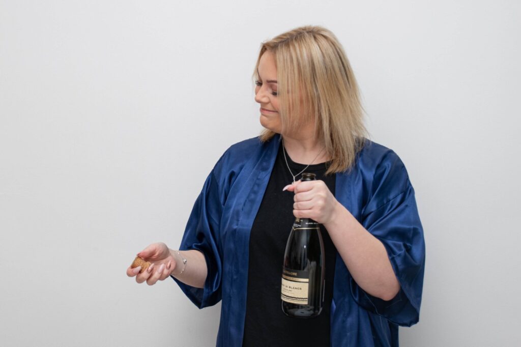 A woman in a blue satin robe holds a bottle of champagne and a cork, smiling as she prepares to celebrate.