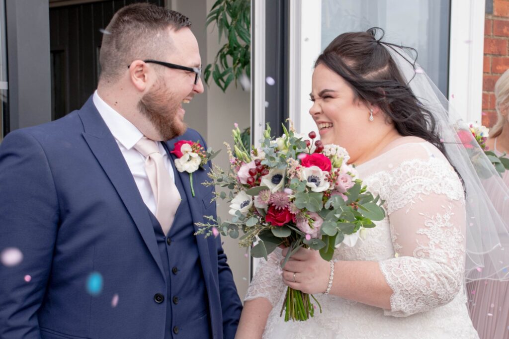A smiling couple shares a joyful moment outdoors, surrounded by colorful confetti, as the bride holds a vibrant bouquet.